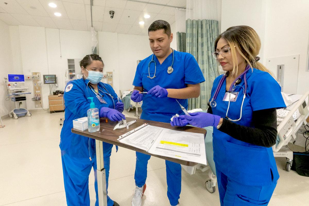 Three students wearing scrubs, working in a hospital room.